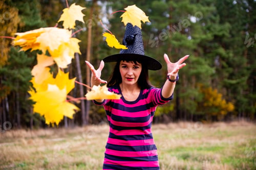 Preview: Beautiful smily witch throw autumn leaves in autumn forest. Halloween celebration party concept