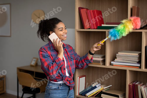 Preview: Smiling millennial african american lady dusting and calling by phone, enjoy household chores in