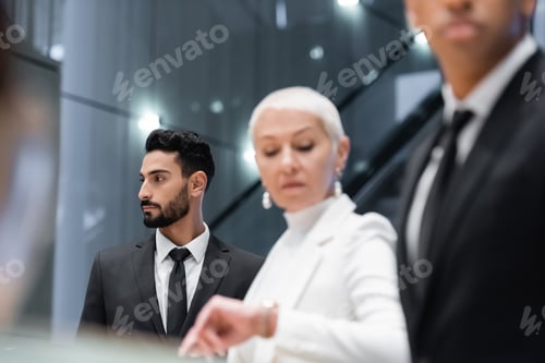 Preview: bi-racial security man near senior businesswoman checking time in hotel on blurred foreground