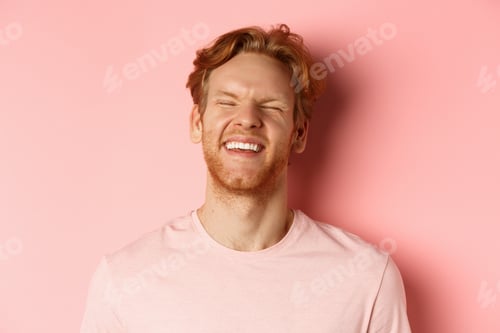 Preview: Close up of carefree man with red hair and beard, laughing and feeling happy, standing over pink
