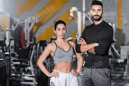 Preview: Young middle east sportswoman looking at camera near arabian friend in gym