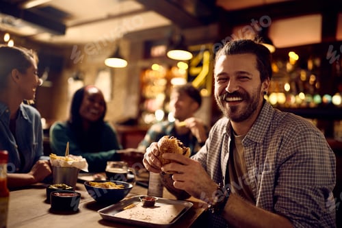 Preview: Happy man eating hamburger in a pub and looking at camera.