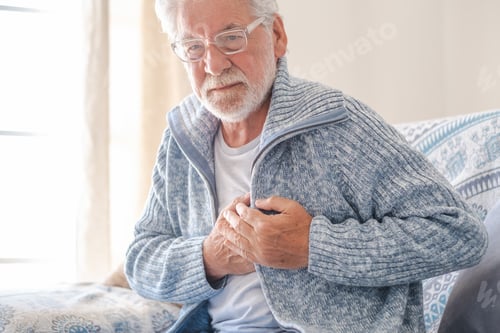 Preview: Worried 70s senior man in light blue sweater sitting at home on sofa suffering from bad chest pain