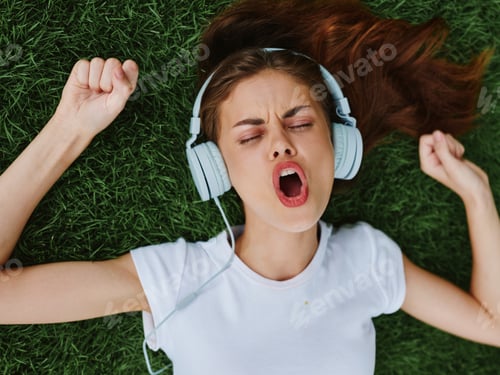 Preview: Beautiful woman in headphones listening to music lying on the grass green lawn in the park in the