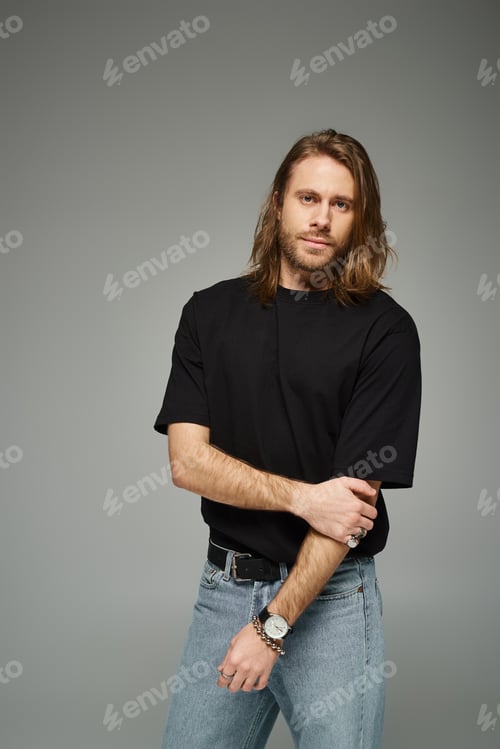 Preview: handsome bearded man with long hair standing in casual t-shirt and jeans on grey background