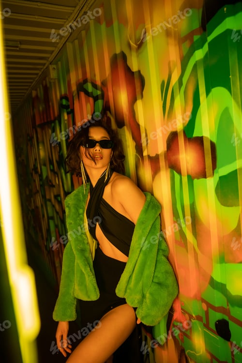 Preview: Sexy asian woman in sunglasses and fake fur jacket standing near graffiti in night club