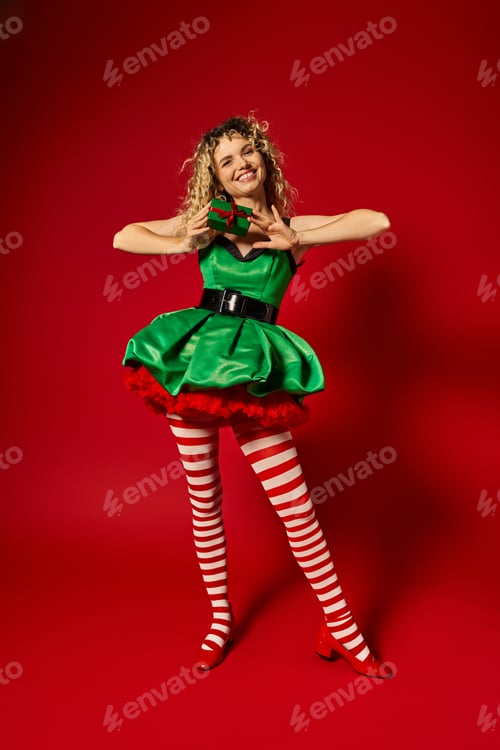 Preview: attractive woman dressed as new year elf smiling and holding present at camera on red backdrop
