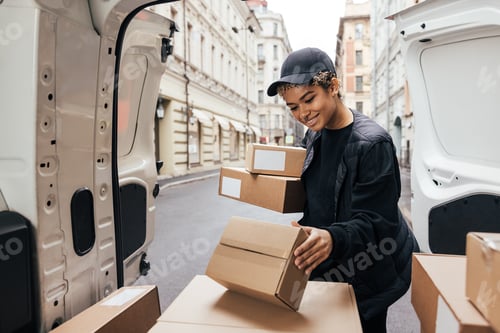 Preview: Smiling woman courier in uniform standing at car trunk holding boxes