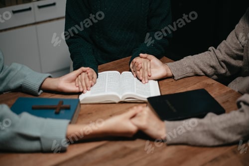 Preview: Group of Christians sit together and pray around a wooden table with blurred open Bible pages