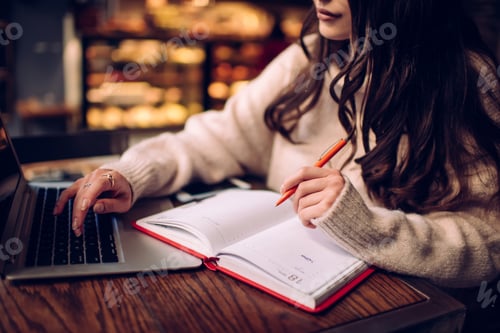 Preview: Crop focused young lady taking notes in daily planner while using computer in modern cafe