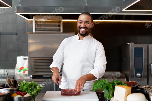 Preview: Handsome young African chef standing in professional kitchen in restaurant preparing a meal of meat