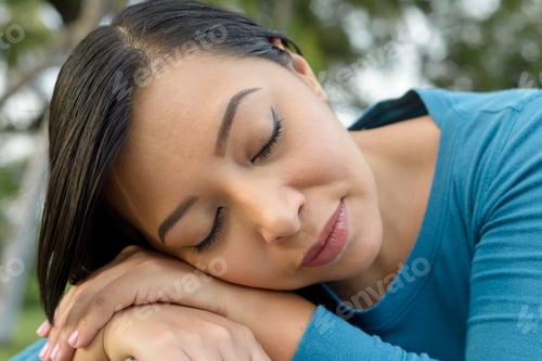 Preview: Woman with dark hair and a blue shirt is sleeping with her head resting on her arms.