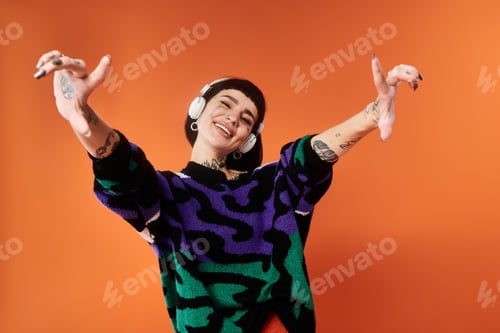 Preview: Young woman with vibrant tattoos and short hair posing in a bright sweater against orange
