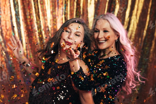 Preview: Two senior girlfriends having fun in studio blowing confetti