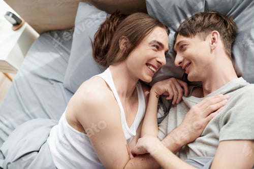 Preview: Top view of cheerful and tattooed homosexual couple in sleepwear talking with each other while lying