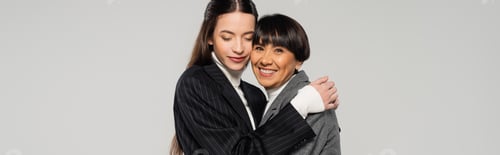 Preview: young woman with closed eyes embracing happy asian mother smiling at camera isolated on grey, banner
