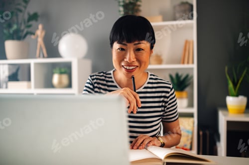 Preview: Smiling asian woman works on laptop with open book in home office