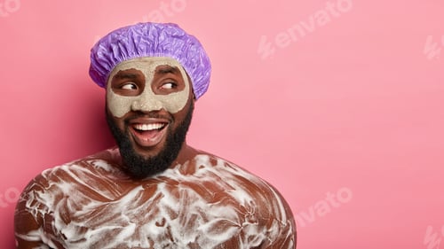 Preview: Horizontal shot of satisfied black man has thick stubble, wears bath hat, clay mask, applies shower