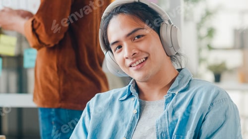 Preview: Young Asian designer man listening to music on headphones, looking at camera and smiling