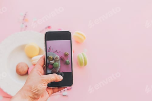 Preview: Hand holding phone and taking photo of stylish colorful macaroons in vintage plate