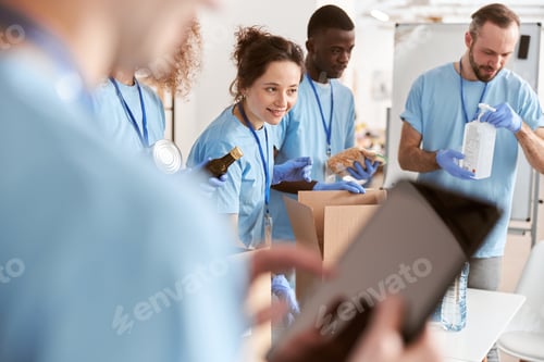 Preview: Diverse volunteers in blue uniform sorting foodstuff in cardboard boxes, working together on