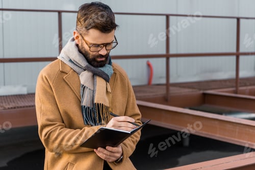 Preview: Bearded businessman standing in coat and writing in notebook