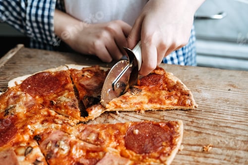 Preview: the hand holds a round knife and cuts a pizza with salami, close-up
