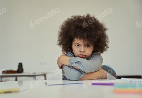 Preview: Disgruntled little schoolboy with afro hair looking resentful, frowning while sitting with arms