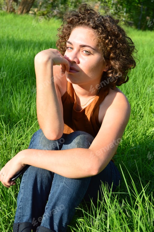 Preview: Vertical shot of a Caucasian young female with light eyes and curly hair sitting on the grass