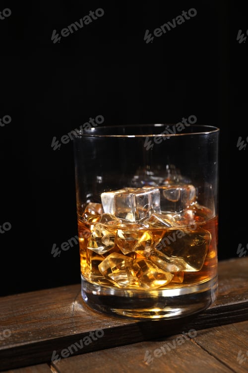 Preview: Whiskey with ice cubes in glass on wooden crate against black background, closeup