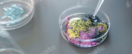 Preview: Drop of liquid falling from from pipette over petri dish with mix of colorful glitters in laboratory