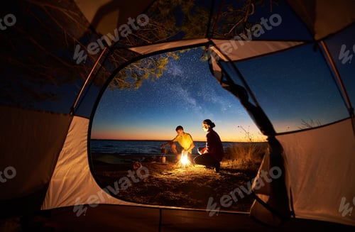 Preview: Couple man and woman having rest at tourist tent and burning campfire on sea shore near forest
