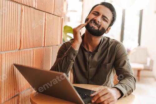 Preview: Middle aged Indian freelancer with closed eyes experiencing toothache working on laptop in cafe