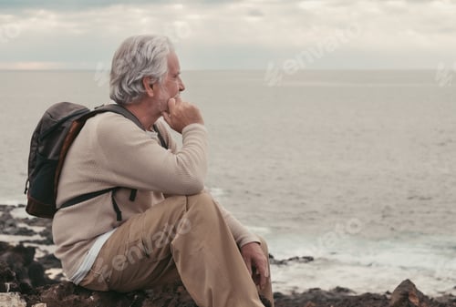 Preview: Pensive mature adult elderly man sitting on the cliff on excursion into the sea at sunset
