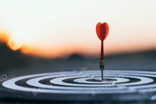Preview: Close up red dart arrow hitting target center dartboard on sunset background.