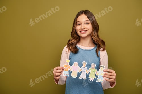 Preview: Smiling preteen girl in denim sundress holding drawn paper characters and looking at camera