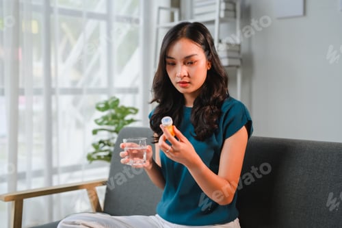 Preview: Young woman taking medicine at home sitting on the sofa