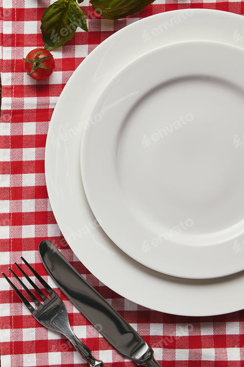 Preview: top view of empty white plates, cutlery and fresh basil with tomato on checkered tablecloth