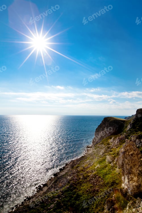 Preview: Seascape of still blue sea waters shore, rocky coastline and bright sun