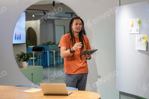 Preview: African American man presenting with tablet, gesturing at office table with laptop and whiteboard