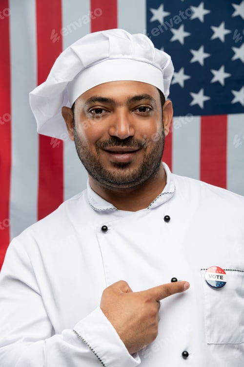 Preview: Smiling male chef of restaurant in uniform pointing at vote insignia on chest