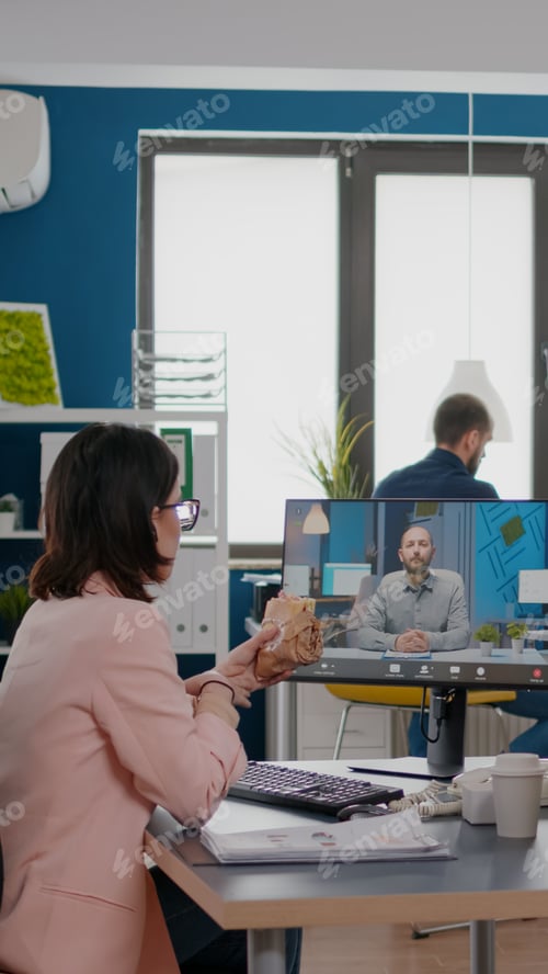Preview: Entrepreneur woman sitting at desk in company office eating sandwich during online videocall