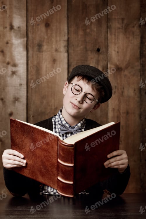 Preview: Young boy in retro clothing wearing spectacles reading a massive book thooughtfully