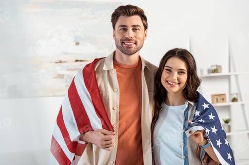 Preview: Selective focus of smiling couple looking at camera while wrapping in american flag