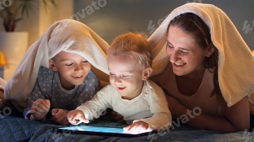 Preview: Happy smiling kids with mother lying under blanket and using tablet computer