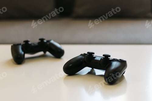 Preview: Photo of two gamepads with selective focus, on white table with gray sofa in the background