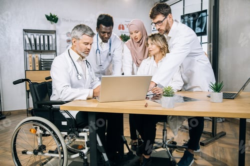 Preview: Aged doctor in wheelchair using laptop with diverse partners