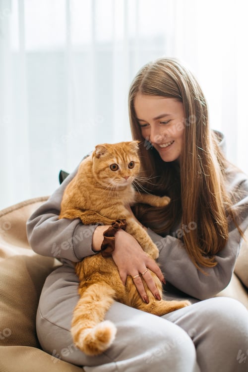 Preview: Young woman relaxing on sofa holding her ginger cat