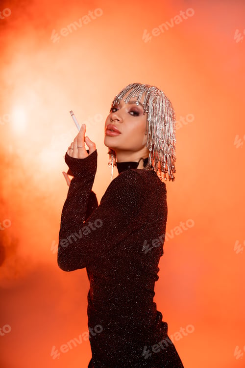 Preview: glamour woman in black dress and shiny headwear looking at camera while posing with cigarette on