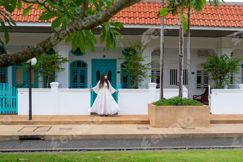 Preview: girl in white dress stands against the background of a white building with blue shutters and a fence